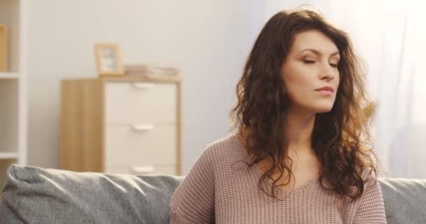 Retrato de la hermosa joven con el pelo rizado girando la cabeza hacia la cámara y sonriendo en casa en el sofá. En el interior. De cerca. — Vídeos de Stock