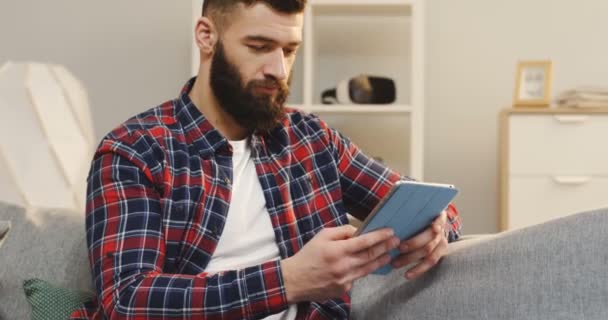 Bel giovanotto con la camicia variopinta e con la barba che lavora sul tablet mentre è seduto sul divano nell'accogliente stanza. Dentro. — Video Stock