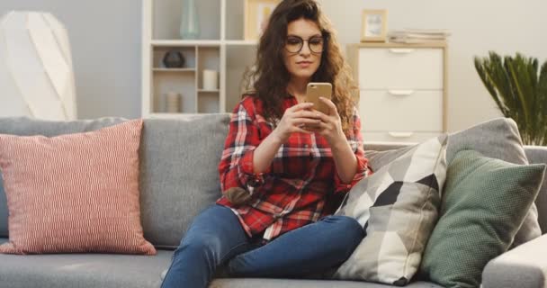 Красивая молодая женщина в очках, заклеивающая смартфон и улыбающаяся, сидя дома на диване. В помещении — стоковое видео