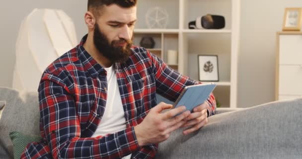 Bonito jovem na camisa xadrez e com uma fita de barba no dispositivo tablet enquanto sentado no sofá na acolhedora sala de estar. Interior — Vídeo de Stock