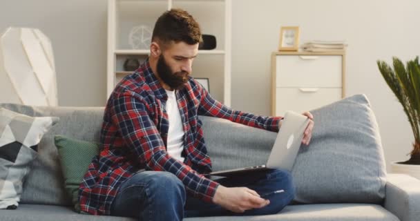 Homem bonito com a camisa xadrez trabalhando no computador portátil e falando ao telefone na sala de estar. Interior — Vídeo de Stock