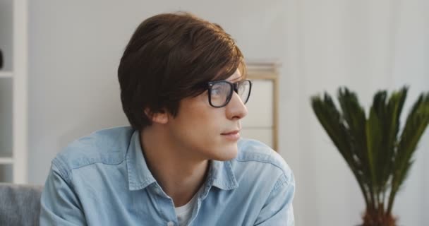 Primer plano del joven atractivo en gafas girando la cabeza y sonriendo sinceramente a la cámara en la habitación moderna del apartamento. Adentro. Retrato — Vídeo de stock