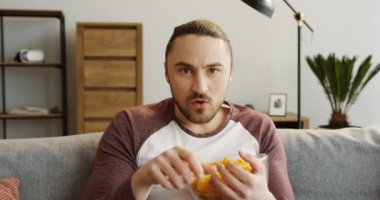 Koltukta oturan, Tv izlerken ve rahat oturma odasında cipsi yemek genç beyaz adam portresi. Yakın çekim. Kapalı