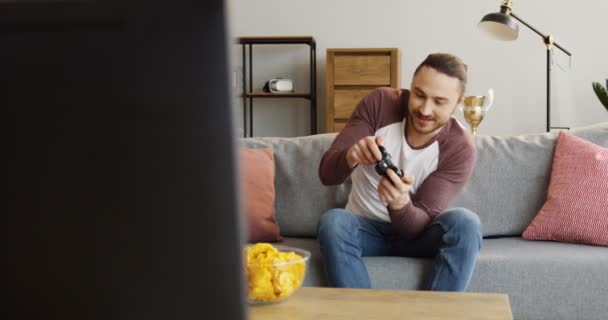 Молодой кавказский мужчина сидит на диване в уютной комнате и играет в видеоигры, а потом нервничает, потому что проигрывает. В помещении — стоковое видео