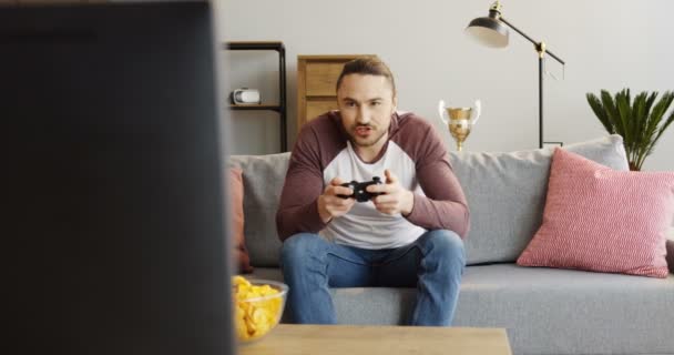 若いハンサムな男は居間のソファに座って、ビデオ ゲームをプレイします。中に — ストック動画