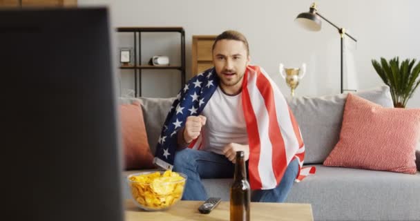 Giovane uomo attraente coperto con bandiera americana seduto sul divano, guardando la partita sportiva e ottenere felice di vincere. Interni — Video Stock