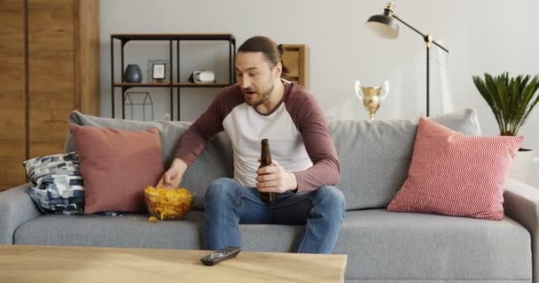 Kaukasische goed uitziende man kijken naar sport spel, chips eten en het drinken van bier. Wordt nerveus. Thuis. Binnenshuis — Stockvideo