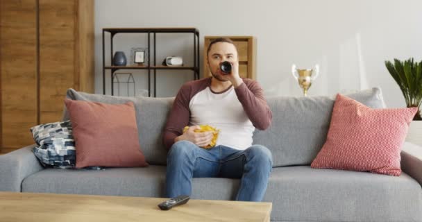 Jeune homme beau se reposant devant la télévision tout en regardant le jeu de sport, manger des chips et boire de la bière dans le salon. À l'intérieur — Video