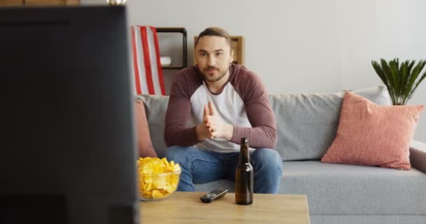 Junger attraktiver Mann, der fernsieht, Chips mit Bier isst und kommentiert, was er sieht. Wohnzimmer mit amerikanischer Flagge dahinter. Innenräume — Stockvideo