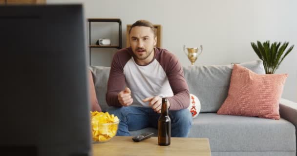 Retrato tiro do homem caucasiano animando-se enquanto assiste jogo desportivo na TV em casa. Cerveja e batatas fritas. Interior — Vídeo de Stock