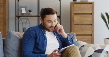 Bir tablet ellerde ile kanepede oturan ve telefonda konuşmayı iyi görünümlü genç adam. İçinde