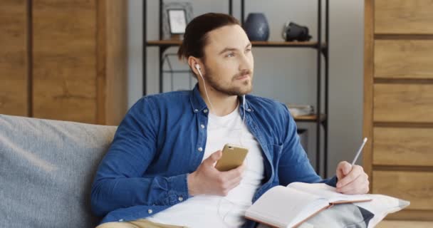 Porträt des jungen kaukasischen Mannes, der über Kopfhörer vom Smartphone Musik hört und sich Notizen im Notizbuch macht. zu Hause. innen — Stockvideo