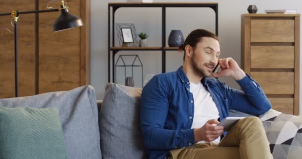 Καυκάσιος νεαρός άνδρας σε casual στυλ κάθεται στον καναπέ με μια συσκευή tablet στα χέρια και να μιλάτε στο τηλέφωνο. Εσωτερική — Αρχείο Βίντεο