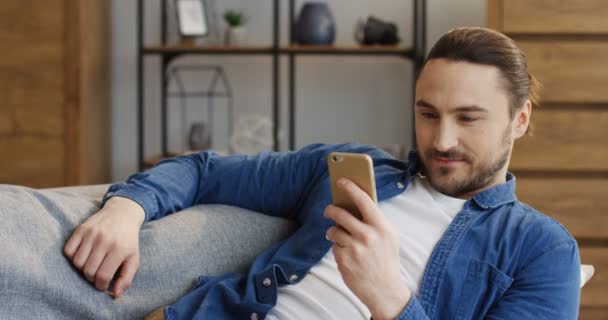 Retrato del atractivo hombre con camisa vaquera descansando en el sofá y enviando mensajes de texto y grabando en el teléfono inteligente en la sala de estar. En interiores — Vídeos de Stock