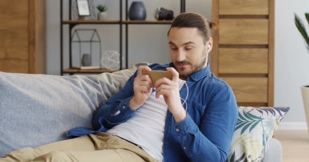 Retrato del joven guapo hombre guapo en los auriculares que yacen en el sofá con almohadas y ver algo en la pantalla del teléfono inteligente horizontalmente. En casa. Adentro. — Vídeo de stock