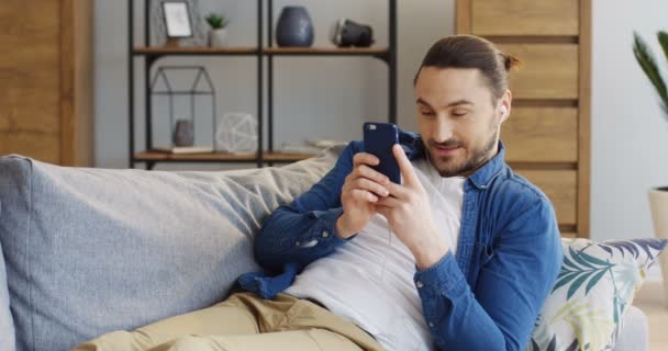Joven hombre atractivo en los auriculares descansando en el sofá con almohadas y riendo mientras ve algo en la pantalla del teléfono inteligente verticalmente. En casa. De interior — Vídeo de stock