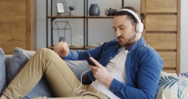 Büyük beyaz kulaklık ve şarkı ve onun elinde smartphone ekranında seyir rahat tarzı yakışıklı genç adam. Rahat oturma odası. İçinde