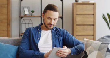 Bir kuyruk ile yastık oturma odasında kanepede oturan ve tablet cihaz üzerinde kayıt kot gömlek ile genç iyi görünümlü adam. Kapalı