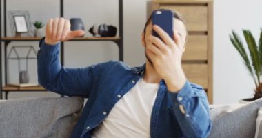 Kot pantolonlu genç beyaz adam kanepede yastıklarla oturuyor ve akıllı telefonda görüntülü konuşma yapıyor. İçeride.