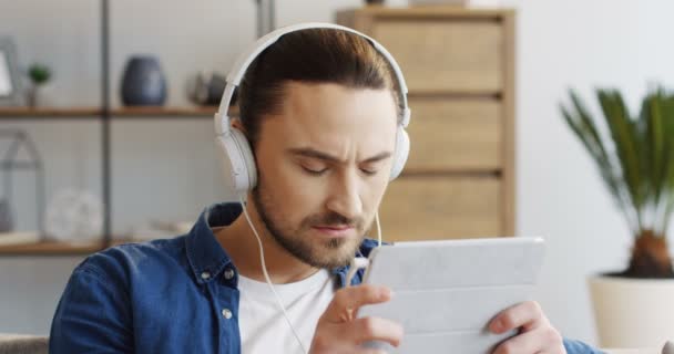 タブレット デバイスで真剣に何かを見ている大きな白いヘッドフォンで若い魅力的な男のクローズ アップ。家に。肖像画。中に — ストック動画