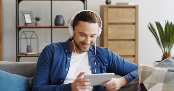 Retrato de la foto del joven atractivo en los grandes auriculares blancos viendo algo divertido en la tableta y riendo. En casa. De interior — Vídeos de Stock