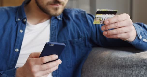Крупный план молодого человека в джинсовой рубашке и покупки в Интернете на смартфоне с кредитной картой в руках. В помещении — стоковое видео