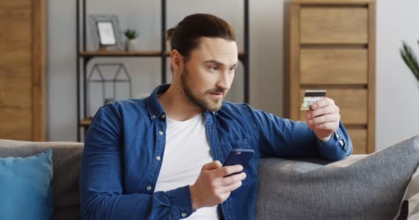 Όμορφος Καυκάσιος άντρας κρατάει ένα smartphone και εισάγοντας τον αριθμό της πιστωτικής κάρτας, ενώ τα ηλεκτρονικά ψώνια. Στο εσωτερικό — Αρχείο Βίντεο