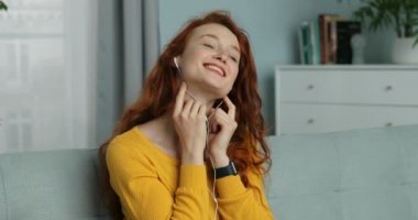 Evdeki kanepede oturan, kızıl saçlı, oldukça neşeli kulaklıklı, genç, beyaz bir kadını yakından çek. Güzel kız akıllı telefondan müzik dinliyor, şarkı söylüyor ve gülümsüyor. Kadın şarkıdan zevk alıyor..