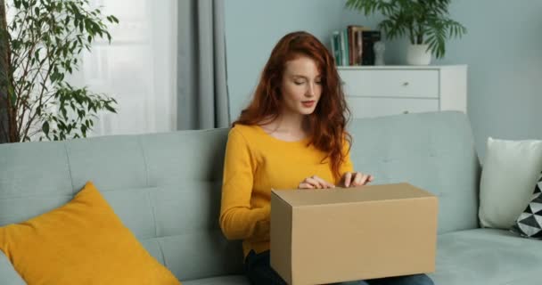 Mladá běloška sedí na gauči v obýváku a otvírá kartónovou krabici. Červenovlasá atraktivní veselá dívka se balíkem doma. Doručení pošty do domu. — Stock video