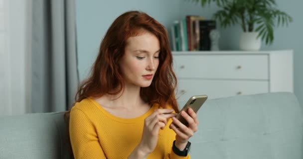 Närbild av kaukasiska unga rödhåriga kvinna sitter på soffan i vardagsrummet och handla online på smartphone. Söt kvinnlig shoppare köper på telefon och betalar med kreditkort. Självisolering i hemmet — Stockvideo