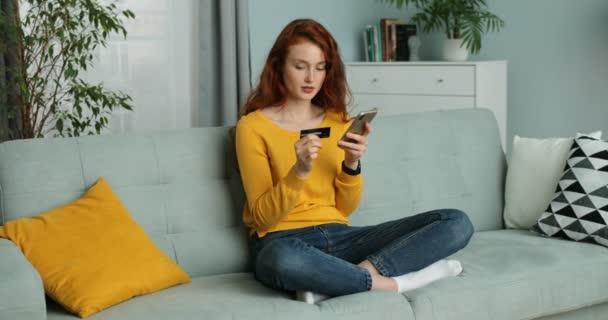 Caucasian young beautiful red-haired woman sitting on couch in living room and shopping online on mobile phone. Pretty girl buying on smartphone and paying with credit card. At home. Self-isolation. — Stock Video