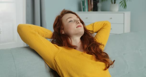 Mujer caucásica joven pelirroja en suéter amarillo sentada en el sofá y pensando mientras descansa en la acogedora sala de estar. Chica bonita descansando en el sofá en casa. Concepto de autoaislamiento. Tiempo de cuarentena . — Vídeos de Stock