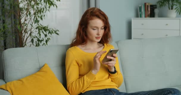 Улыбающаяся молодая белая женщина с рыжими кудрявыми волосами, смс-ками на смартфоне и сидящая дома на диване. Красавица прослушивает и прокручивает на мобильном телефоне в гостиной Селф-изолированная девушка онлайн — стоковое видео