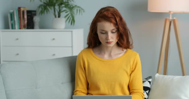 Mujer joven seria caucásica con el pelo largo y rojo sentado en el sofá en casa y escribiendo en el teclado. Hermosa freelancer femenina que trabaja en el ordenador portátil en acogedora sala de estar . — Vídeo de stock