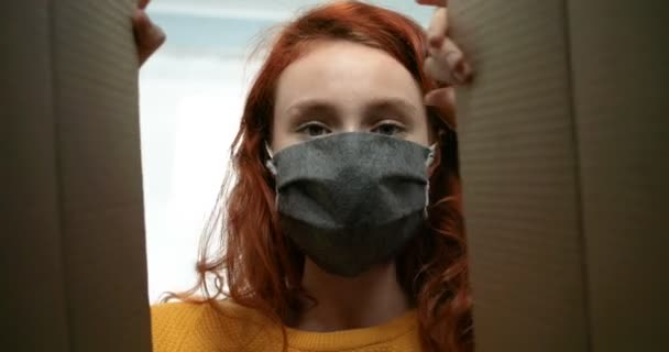 Uitzicht van binnenuit van kartonnen doos op jonge blanke vrouw met rood haar en met masker die erin kijken. Sluiten van mooi meisje openen kartonnen doos. Vrouwelijk pakketje. — Stockvideo