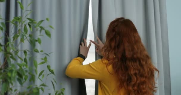 Parte trasera de la mujer joven caucásica con el pelo largo y rizado rojo de pie en la sala de estar en la ventana y cortinas de apertura por la mañana. Vista trasera. Chica comenzando su día Retrato Mujer feliz girando y sonriendo — Vídeos de Stock