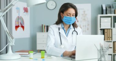 Beyaz elbiseli, mavi maskeli, beyaz saçlı, genç ve güzel bir kadın doktor. Masasında oturmuş, dizüstü bilgisayarında çalışıyor. Klavyede yazan ve hastalık belirtilerini tanımlayan bir kadın doktor.