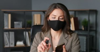 Maskeli güzel beyaz kadın masada oturuyor ve ofisteki peçeteyle akıllı telefonu siliyor. Coronavirus salgını koruması. Kadın temizliği, sterilize etme ve cep telefonunu dezenfektanla dezenfekte etme