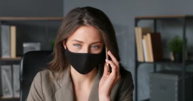 Siyah maskeli güzel beyaz iş kadını ofiste cep telefonuyla konuşuyor. Koronavirüsten korunan güzel bir kadın cep telefonundan konuşuyor. Telefon görüşmesi.
