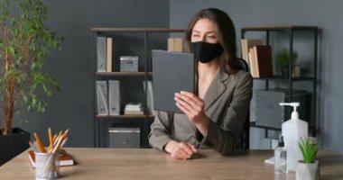 Maskeli şık, beyaz bir kadın ofisinde otururken tablet bilgisayarında video kaset taşıyor. Genç iş kadını web kamerasıyla konuşuyor ve internette görüntülü sohbet yapıyor. Kadın video blogcusu.