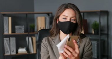 Maskeli güzel beyaz kadın ofis çalışanı masada oturuyor ve akıllı telefonu peçeteyle siliyor. Coronavirus salgını koruması. Kadın temizliği, sterilize etme ve cep telefonunu sterilize etme.