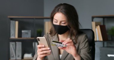 Genç beyaz kadın elinde kredi kartıyla ofiste oturuyor ve akıllı telefondan internetten alışveriş yapıyor. Maskeli güzel bir iş kadını telefonla internet alıyor ve para ödüyor. Karantinada alışveriş yapan kişi.