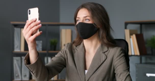 Schöne junge kaukasische Geschäftsfrau in Maske sitzt im Büro und macht Selfie-Fotos mit der Smartphone-Kamera. Hübsche Frau macht Selfies mit dem Handy. Coronavirus-Pandemiekonzept. — Stockvideo