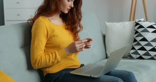 Kaukasisk ung snygg rödhårig kvinna sitter på soffan och handlar online med kreditkort. Internetbutik hemifrån. Söt flicka köper och betalar på soffan i vardagsrummet. — Stockvideo