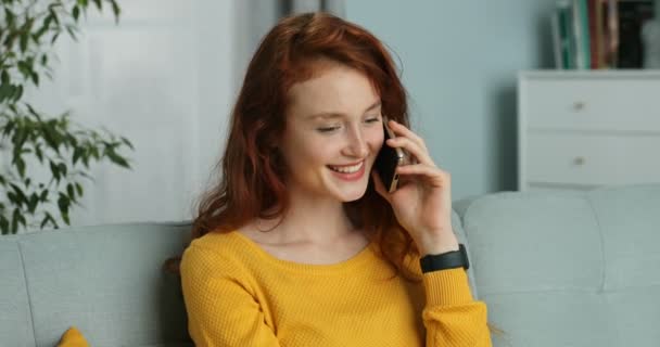 Joyeux jeune femme aux cheveux roux caucasienne parlant sur son téléphone portable et souriant et s'asseyant sur le canapé à la maison. Jolie fille heureuse avec des cheveux bouclés rouges parlant sur téléphone portable dans le salon . — Video