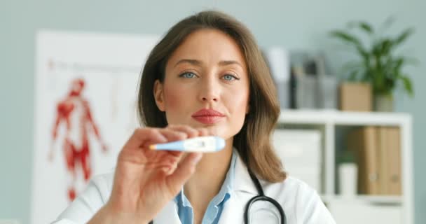 Retrato de una joven mujer caucásica hermosa doctora en bata blanca mostrando termómetro electrónico borroso a la cámara. Médico femenino en su gabinete mostrando alta temperatura como síntoma de enfermedad . — Vídeos de Stock