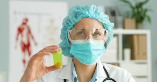 Portrait of young Caucasian woman doctor in white gown, cap, medical mask and goggles showing pills to camera. Female medic demonstrating medicine and cure in tablets. — Stock Video