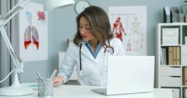 Beyaz önlüklü beyaz kadın doktor dolapta masada oturuyor ve dizüstü bilgisayarda çalışıyor. Klavyede yazı yazan ve hastalık belirtilerini tanımlayan güzel bir kadın..