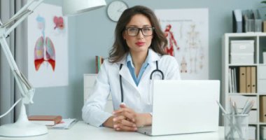 Beyaz cüppeli beyaz kadın doktor portresi rahat bir ofiste masada oturuyor, yüzünü kameraya çevirip mutlu bir şekilde gülümsüyor. Laptop bilgisayarındaki güzel bayan doktor..