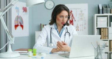 Kabinedeki dizüstü bilgisayardan hastayla konuşan, kulaklıklı beyaz bir bayan doktor. Kadın doktor internet danışmanlığı yapıyor..